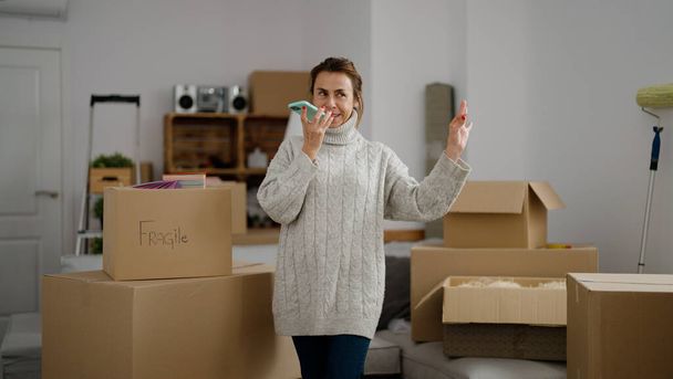 Middelbare leeftijd Spaanse vrouw glimlachend vol vertrouwen praten op de smartphone in het nieuwe huis - Foto, afbeelding