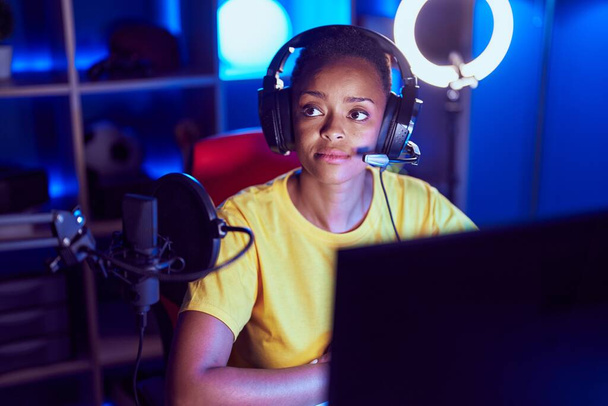 Afrikaans amerikaanse vrouw streamer zitten op tafel met serieuze expressie in gaming kamer - Foto, afbeelding