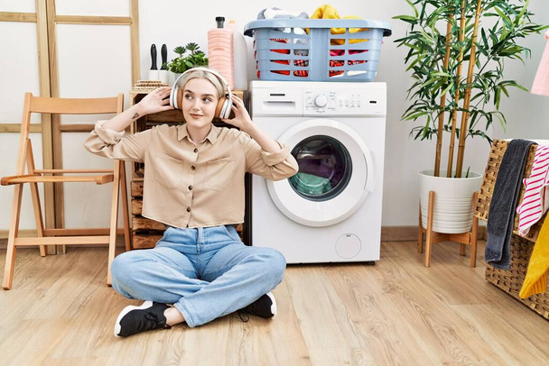 Jovem caucasiana ouvindo música esperando por máquina de lavar roupa na lavanderia - Foto, Imagem