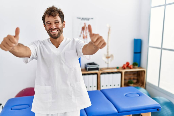 Mladý pohledný fyzioterapeut pracující na klinice pro zotavení bolesti, který dělá pozitivní gesto rukou, palce nahoru, usmívá se a je šťastný z úspěchu. gesto vítěze.  - Fotografie, Obrázek