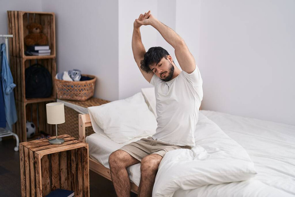 Young hispanic man waking up stretching arms at bedroom - Foto, afbeelding