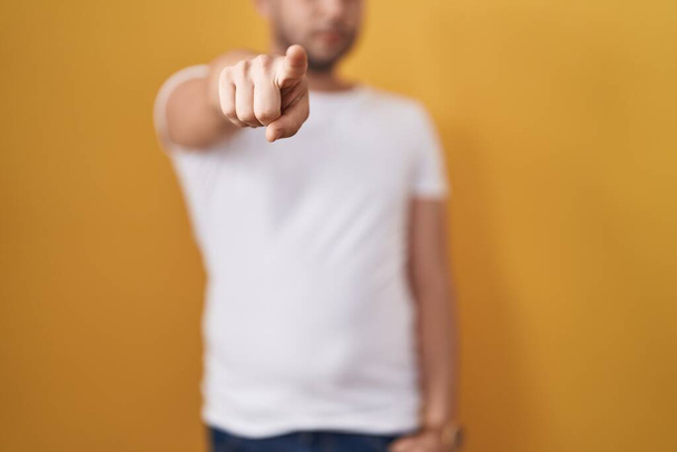 Homme hispanique portant t-shirt blanc sur fond jaune pointant du doigt la caméra et à vous, geste confiant regardant sérieux  - Photo, image