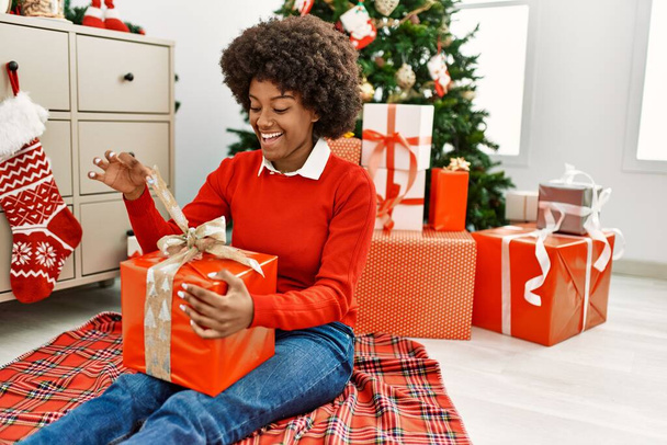 Молодая африканская американка распаковывает подарок сидя у рождественской елки дома - Фото, изображение