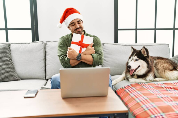 Young hispanic man hugging gift sitting on sofa with dog at home - Foto, afbeelding