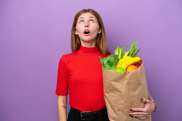 Молодая английская женщина держит сумку с продуктами изолированы на фиолетовом фоне глядя вверх и с удивленным выражением - Фото, изображение