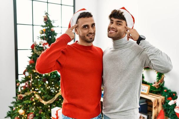 Young gay couple standing by christmas tree wearing hat smiling pointing to head with one finger, great idea or thought, good memory  - Photo, Image