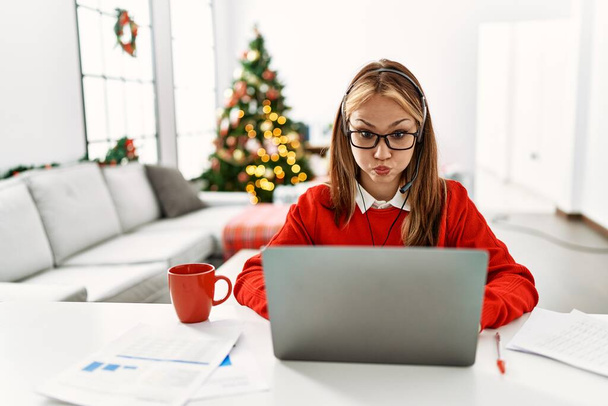 Joven chica caucásica sentada en la mesa de trabajo con el ordenador portátil por el árbol de Navidad hinchando las mejillas con la cara divertida. boca hinchada de aire, expresión loca.  - Foto, Imagen