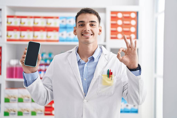 Handsome hispanic man working at pharmacy drugstore showing smartphone screen doing ok sign with fingers, smiling friendly gesturing excellent symbol  - Fotoğraf, Görsel