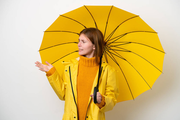 Young English woman with rainproof coat and umbrella isolated on white background with surprise expression while looking side - Photo, image