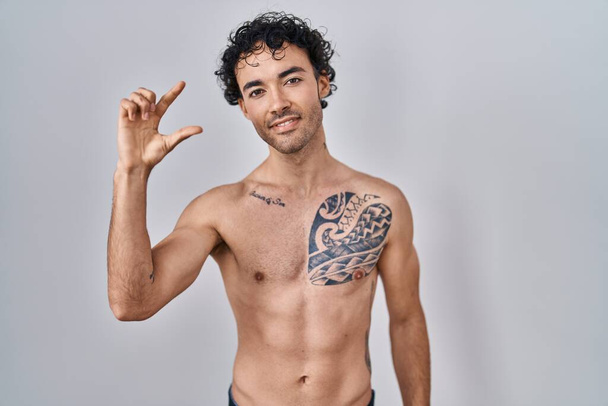 Hispanic man staat shirtloos glimlachend en zelfverzekerd gebaar met de hand doen klein formaat teken met vingers kijken en de camera. meetconcept.  - Foto, afbeelding