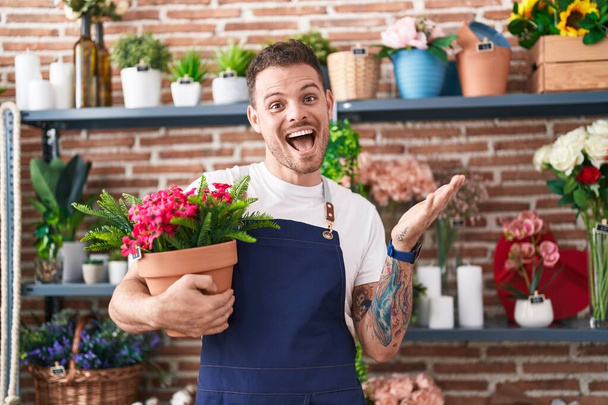 幸せな笑顔で達成を祝う植物ポットを保持する花屋の店で働く若いヒスパニック系の男と手を上げて受賞式  - 写真・画像