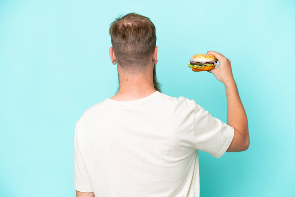 Rotschopf mit langem Bart hält einen Burger isoliert auf blauem Hintergrund in Rückenlage - Foto, Bild