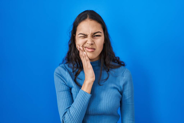 Mavi arka planda duran Brezilyalı genç bir kadın diş ağrısı ya da diş ağrısı yüzünden elleriyle ağıza dokunuyor. dişçi  - Fotoğraf, Görsel
