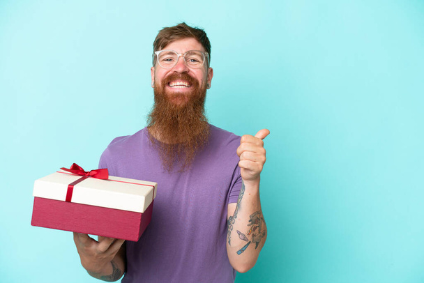 Uzun sakallı kızıl saçlı adam mavi arka planda izole edilmiş bir hediye tutuyor. Ürünü sunmak için yan tarafa bakıyor. - Fotoğraf, Görsel