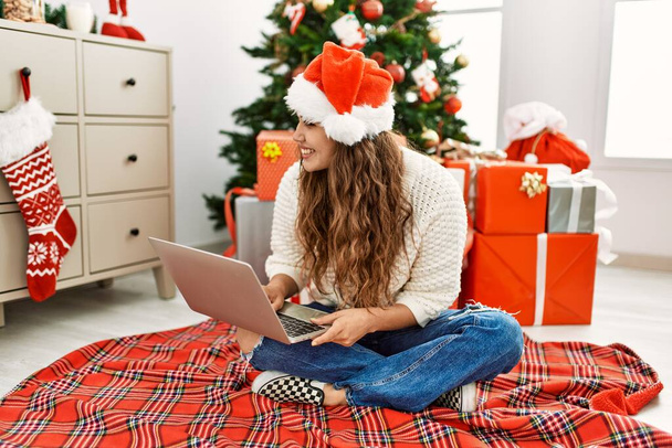 Young beautiful hispanic woman using laptop sitting by christmas tree at home - Fotoğraf, Görsel