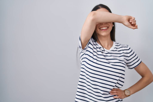 Jeune femme brune portant un t-shirt rayé couvrant les yeux avec le bras souriant gai et drôle. concept aveugle.  - Photo, image