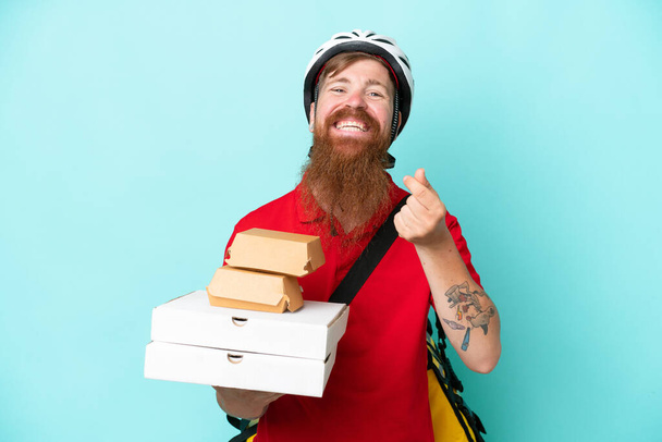 Liefermann hält Pizzen und Burger isoliert auf blauem Hintergrund und macht Geld-Geste - Foto, Bild