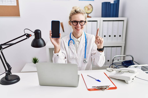 Молодая женщина-врач, держащая смартфон, показывает чистый экран, улыбающийся идеей или вопросом, указывая пальцем со счастливым лицом, номер один  - Фото, изображение