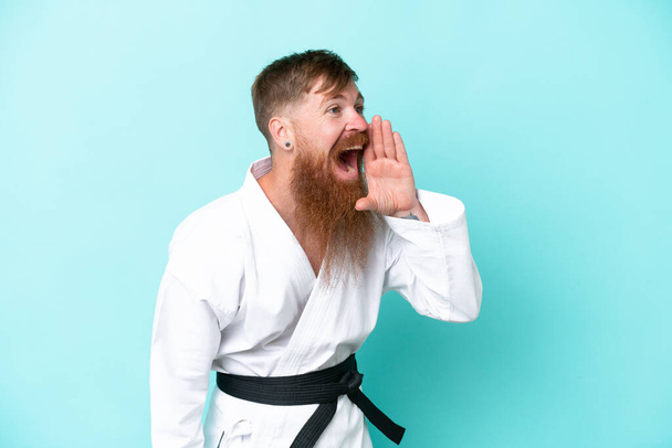Redhead man with long beard doing karate isolated on blue background shouting with mouth wide open to the side - Photo, Image