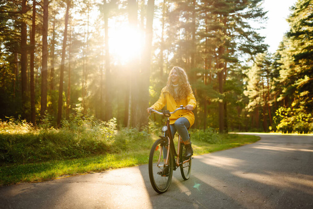 Beautiful happy woman in yellow coat riding bicycle in autumn park. Autumn fashion. Lifestyle. Relax, nature concept. - Photo, image