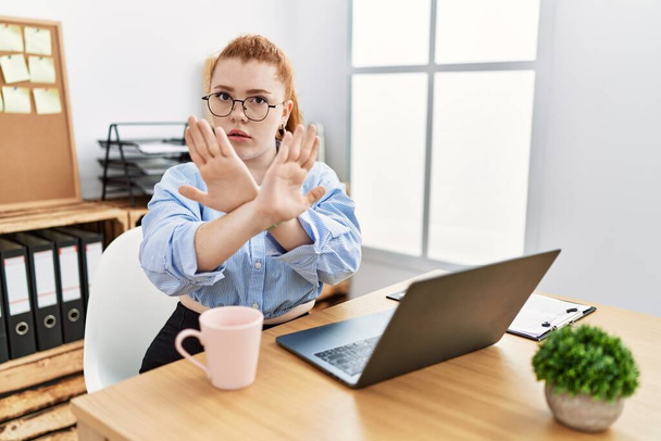Ofiste çalışan genç kızıl saçlı bir kadın bilgisayardan reddedilme ifadesi kullanıyor. Kollarını kavuşturuyor ve avuç içleri negatif işaretlerle dolu. Öfkeli bir yüz.  - Fotoğraf, Görsel