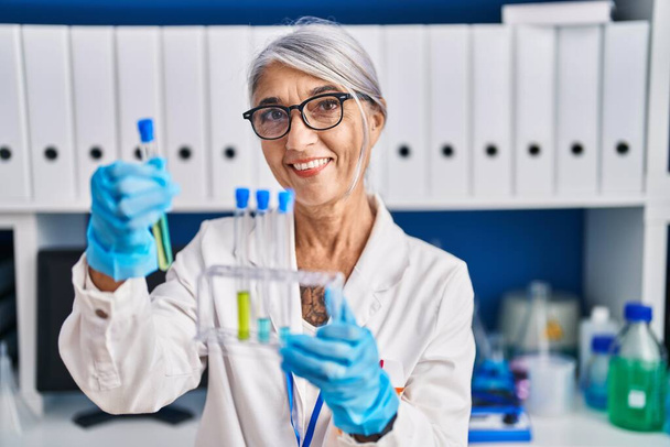 Middle age grey-haired woman scientist smiling confident holding test tubes at laboratory - Фото, изображение