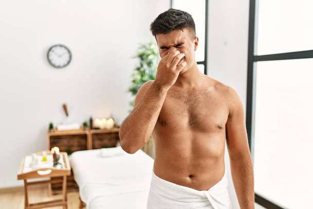 Young hispanic man standing shirtless at spa center smelling something stinky and disgusting, intolerable smell, holding breath with fingers on nose. bad smell  - Photo, Image