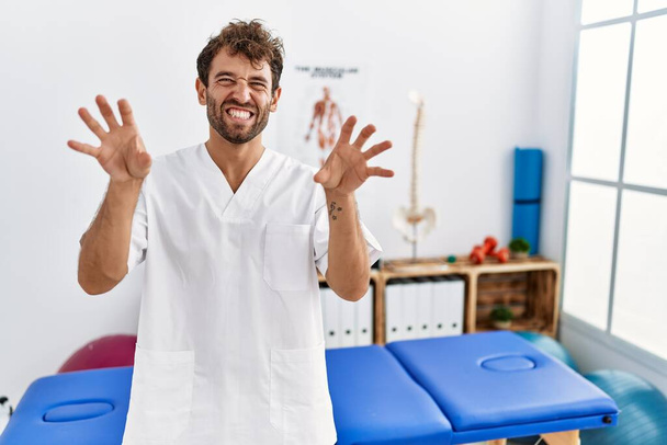 Jonge knappe fysiotherapeut man werken bij pijn herstel kliniek lachen grappig doen klauw gebaar als kat, agressieve en sexy uitdrukking  - Foto, afbeelding