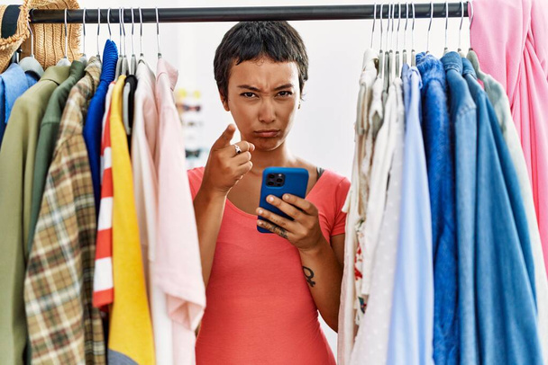 Молода іспаномовна жінка з коротким волоссям шукає одяг на стійці для одягу, використовуючи смартфон, вказуючи пальцем на камеру і до вас, впевнений жест виглядає серйозно
  - Фото, зображення