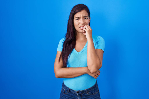 Mujer hispana joven de pie sobre fondo azul mirando estresada y nerviosa con las manos en la boca mordiendo las uñas. problema de ansiedad.  - Foto, Imagen