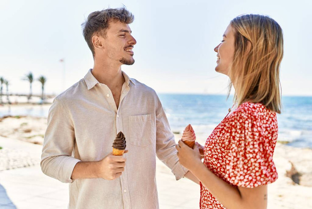 Joven pareja hispana de vacaciones sonriendo feliz comiendo helado en la playa - Foto, imagen