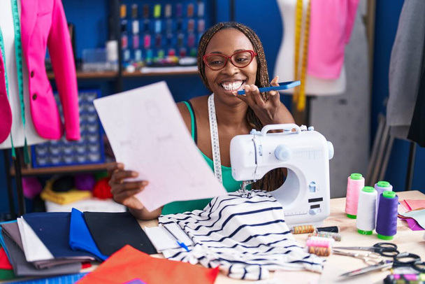 Африканская американка портной говорит на смартфоне глядя дизайн одежды в ателье - Фото, изображение