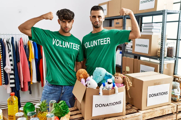 Young gay couple wearing volunteer t shirt at donations stand strong person showing arm muscle, confident and proud of power  - Photo, Image