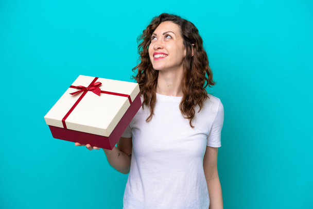 Elinde mavi arka planda izole edilmiş bir hediye tutan beyaz bir kadın gülümserken yukarı bakıyor. - Fotoğraf, Görsel