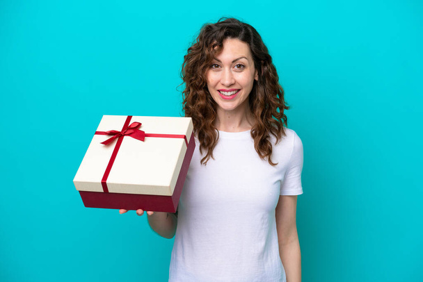 Young caucasian woman holding a gift isolated on blue background with surprise and shocked facial expression - Fotó, kép