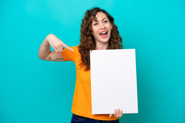 Young caucasian woman isolated on blue background holding an empty placard with happy expression and pointing it - Photo, Image