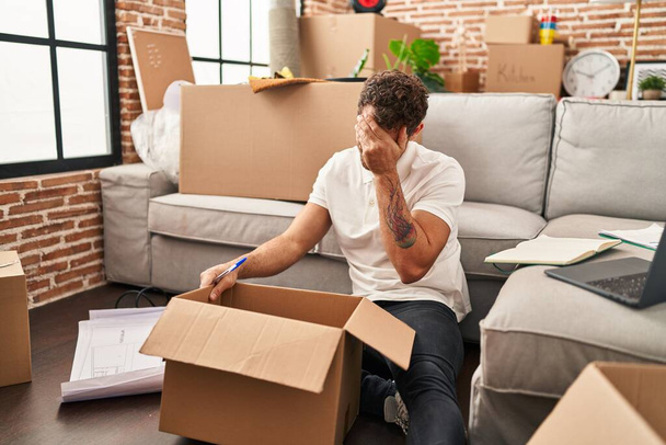 Young hispanic man unboxing package with worry expression at new home - Fotó, kép