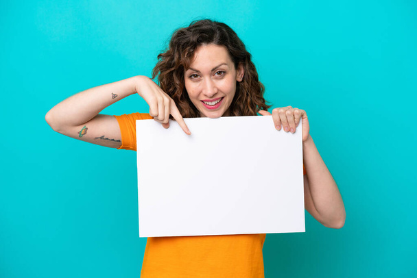 Молодая белая женщина на голубом фоне держит пустой плакат со счастливым выражением лица и направляет его - Фото, изображение