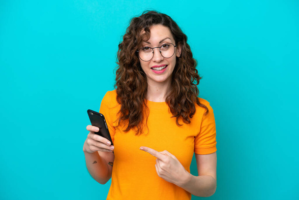 Young caucasian woman isolated on blue background using mobile phone and pointing it - Photo, Image