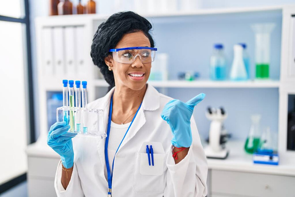 Mujer hispana de mediana edad trabajando en laboratorio científico apuntando con el pulgar hacia el costado sonriendo feliz con la boca abierta  - Foto, Imagen