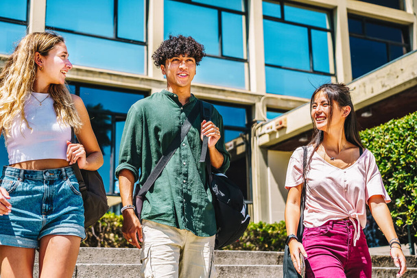Από τα κάτω χαρούμενος πολυφυλετικές φίλους σε casual ρούχα χαμογελώντας και μιλώντας, ενώ το περπάτημα σε βήματα έξω από το σύγχρονο πανεπιστημιακό κτίριο την ηλιόλουστη μέρα - Φωτογραφία, εικόνα