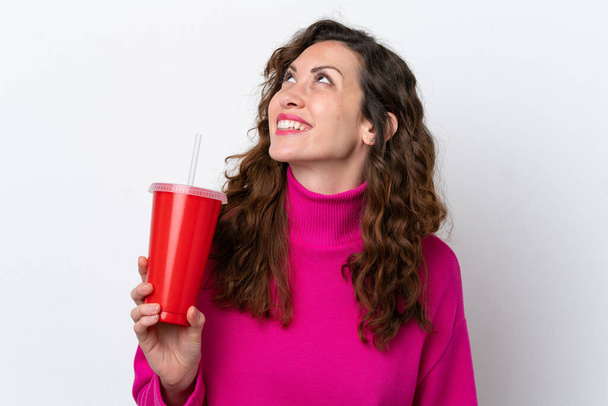 Joven mujer caucásica bebiendo refresco aislado sobre fondo blanco mirando hacia arriba mientras sonríe - Foto, imagen