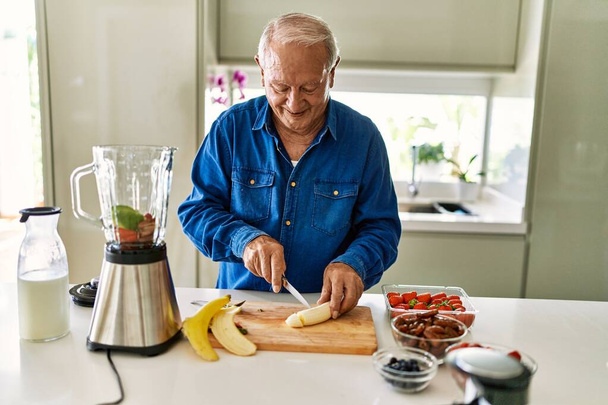 Senior man smiling confident cutting banana at kitchen - Fotoğraf, Görsel