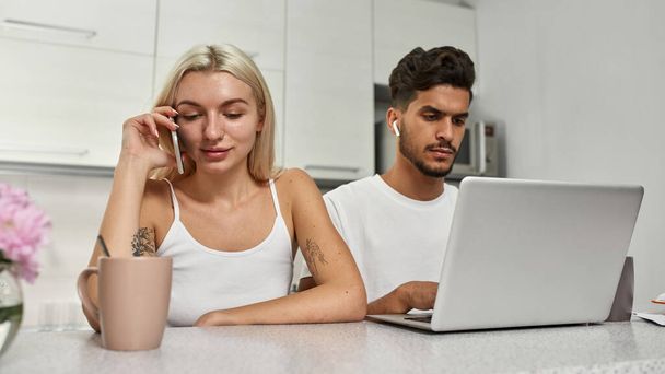 Focused middle eastern man working on laptop while european woman talking on smartphone at table at home kitchen. Young multiracial couple. Remote work and freelance. Domestic lifestyle. Spacious flat - Фото, изображение