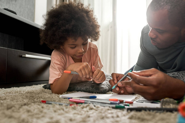 Partial adult father and little son draw with felt tip pens in sketchbook on carpet at home. Young black family spend time together. Fatherhood and parenting. Domestic leisure, hobby and entertainment - Foto, immagini
