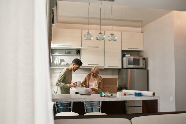 Blonde caucasian girl cutting air bubble film with scissors in hands of middle eastern man at kitchen. Young multiethnic couple packing things for moving to new apartment. Home relocation - Foto, Bild