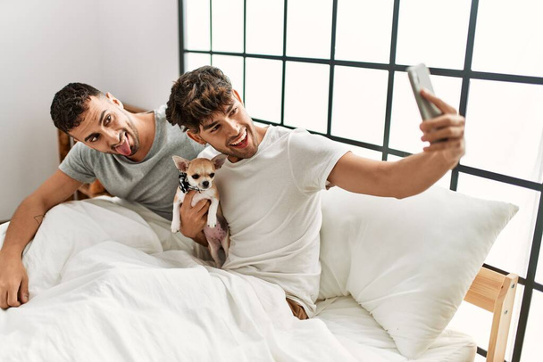Dva hispánští muži pár dělat selfie u smartphonu sedí na posteli s čivava v ložnici - Fotografie, Obrázek
