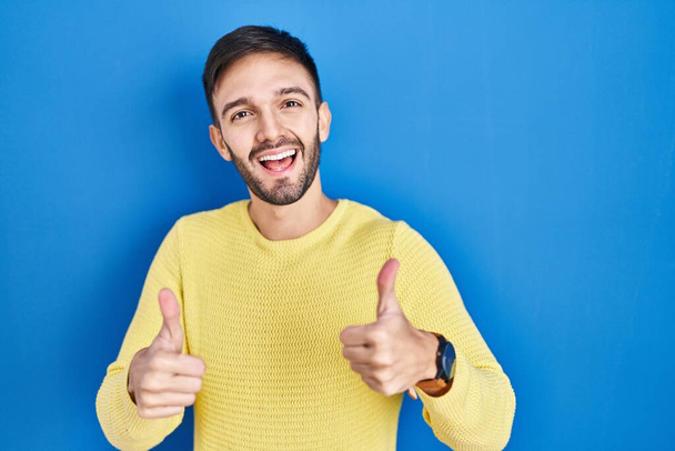Ein spanischer Mann, der über einem blauen Erfolgsschild steht, macht eine positive Geste mit der Hand, Daumen hoch lächelnd und glücklich. Fröhlicher Ausdruck und Siegergeste.  - Foto, Bild