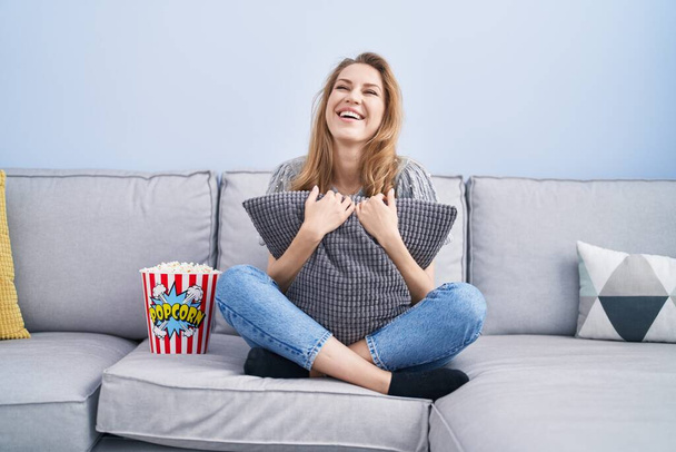 Beautiful blonde woman watching tv sitting on the sofa smiling and laughing hard out loud because funny crazy joke.  - Photo, Image