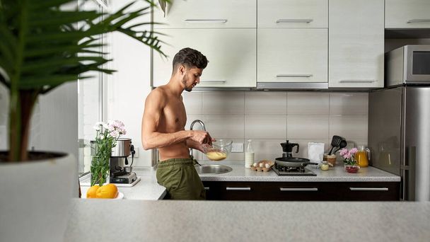 Side view of middle eastern man mixing eggs in bowl with whisk at home kitchen. Young bearded handsome guy with naked torso cooking. Domestic lifestyle. Modern spacious apartment. Morning time - Fotografie, Obrázek